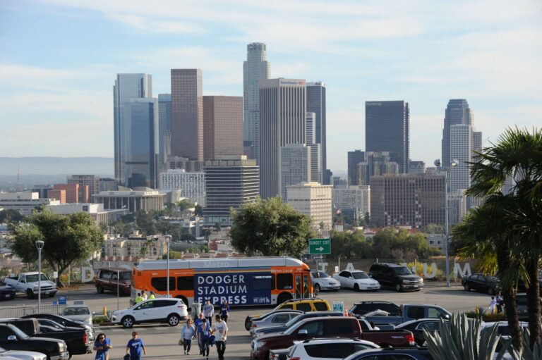 Dodger Stadium Express shuttle bus in foreground with passengers walking towards stadium with Downtown Los Angeles tall buildings in the background.